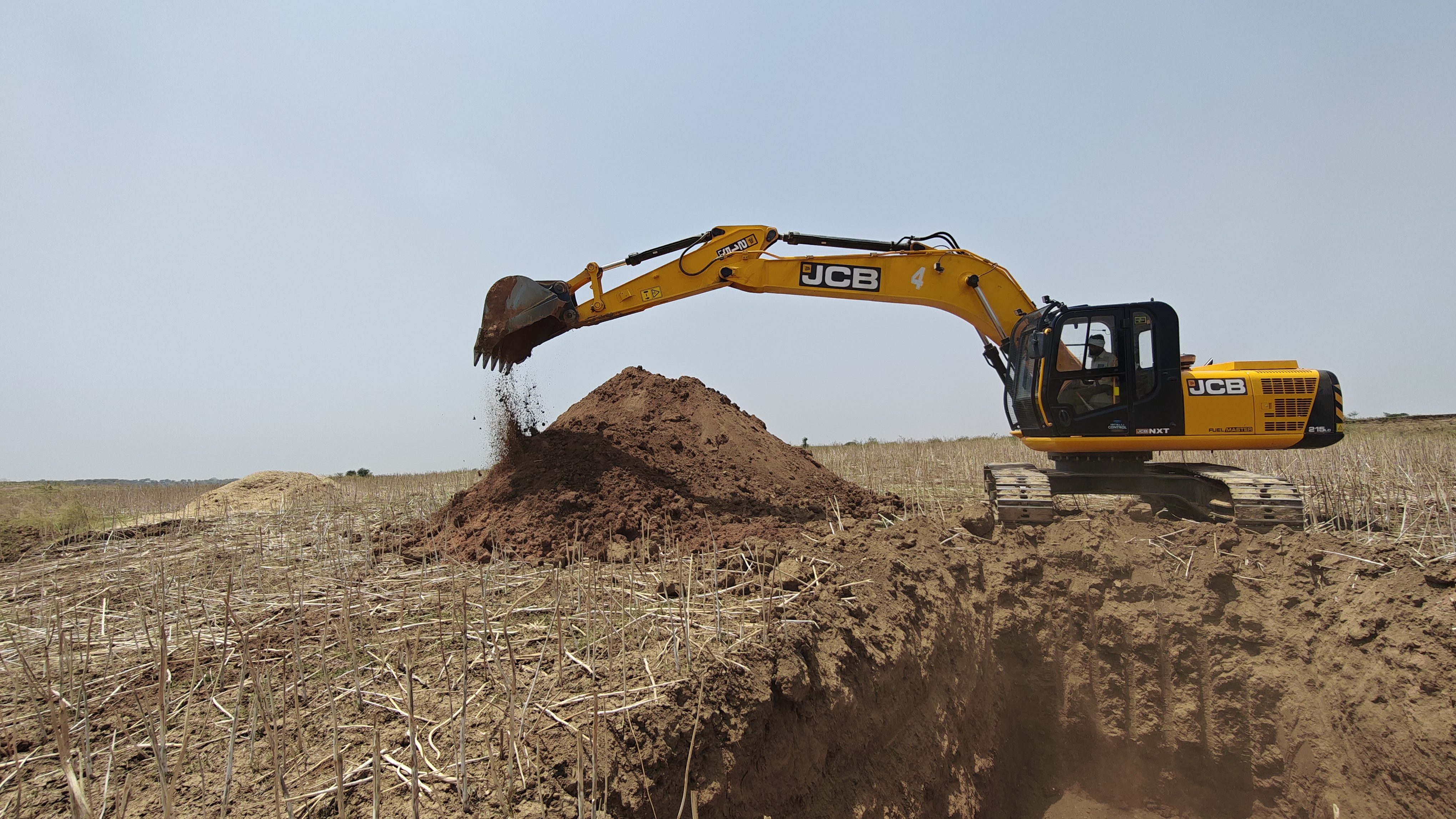 JCB 215 Fuel Master performing soil excavation and earthmoving work in an open field. The machine is being used for land development and excavation activities, showcasing efficient performance in bulk earthwork operations.